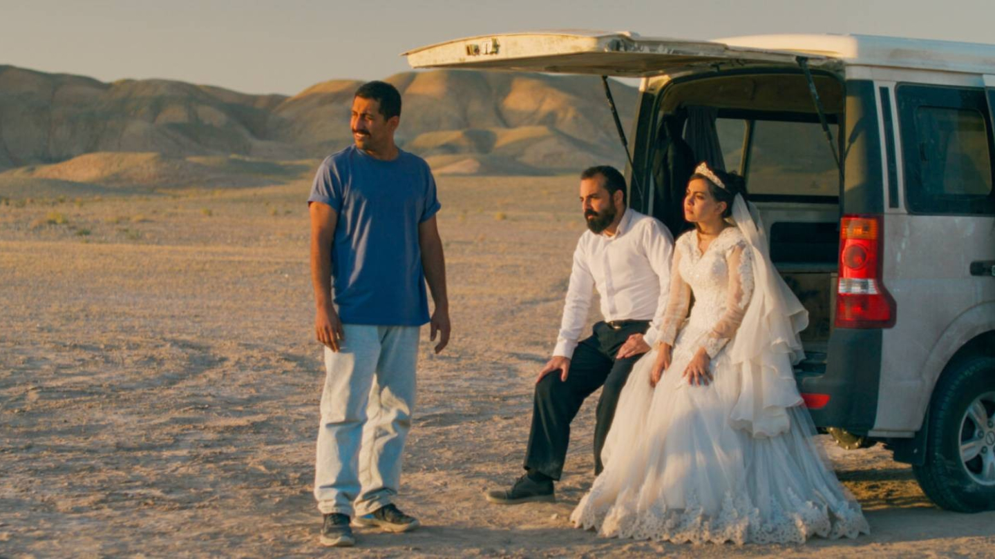 Uma noiva e um homem de camisa social estão sentados no porta-malas aberto de uma van branca quebrada no meio de um deserto, enquanto outro homem, vestindo camiseta azul, observa a paisagem ao lado deles