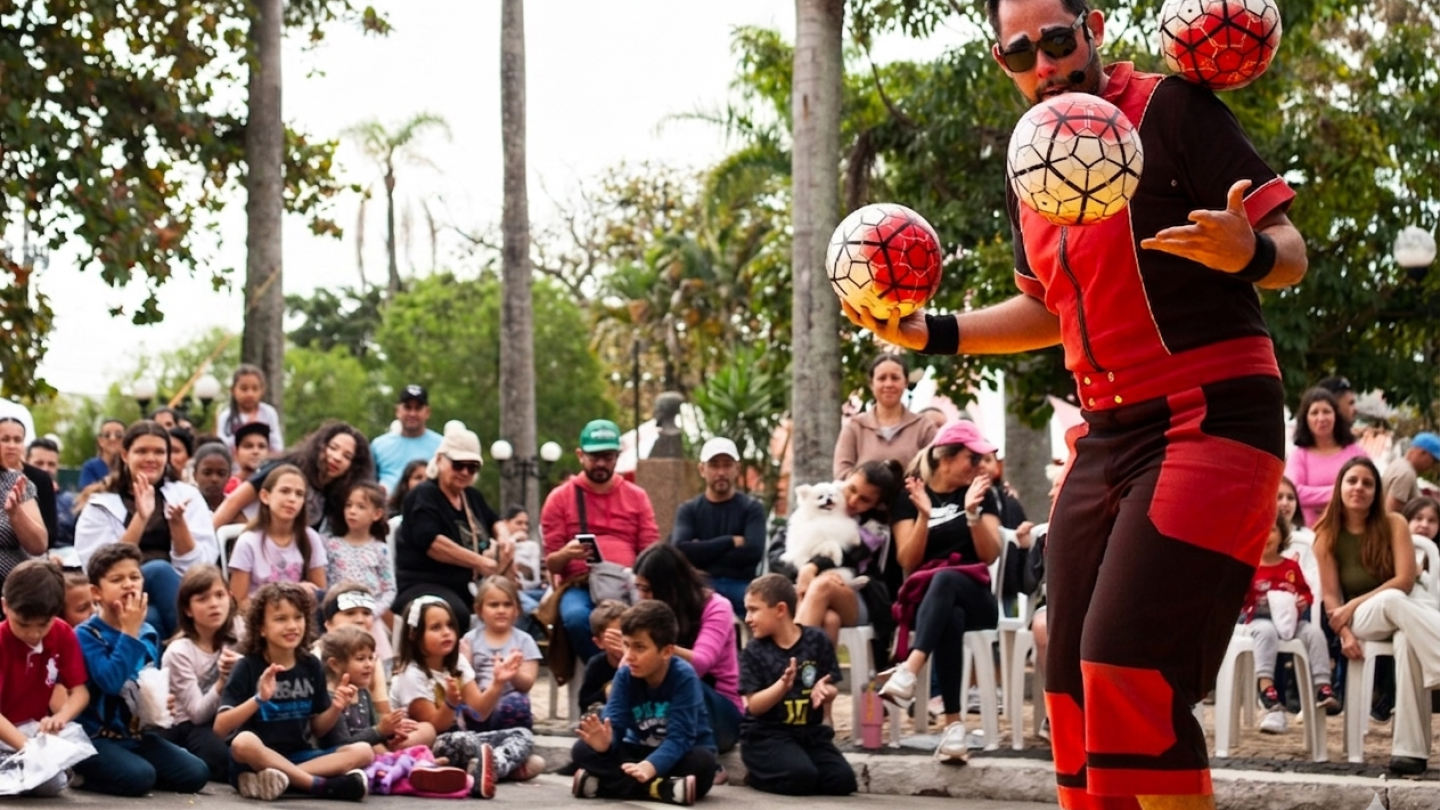 Um artista de rua faz malabarismo com três bolas decoradas diante de uma plateia atenta e sorridente em uma praça pública