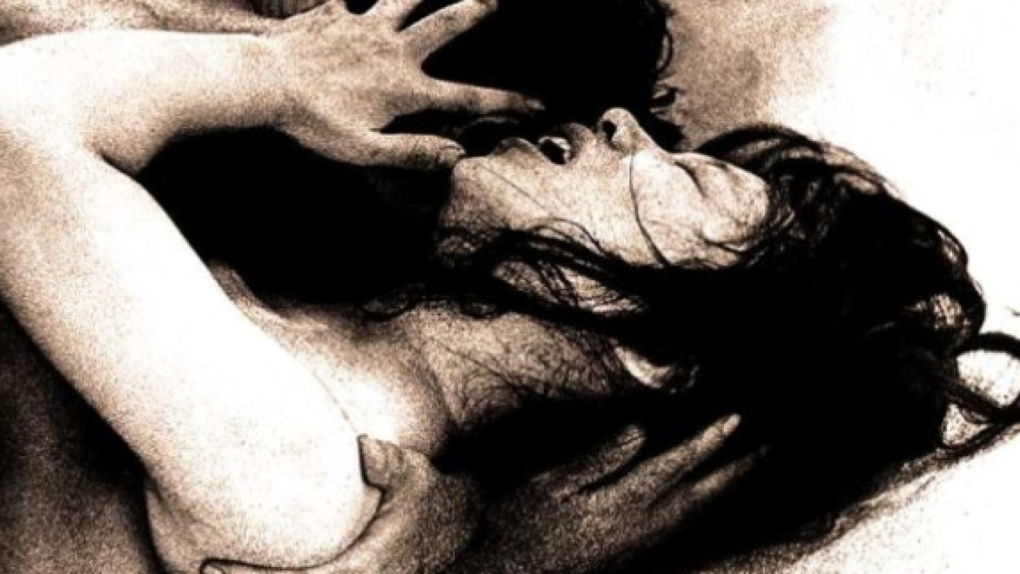 Imagem em preto e branco, de forte caráter artístico e expressivo, retratando duas pessoas em um abraço intenso. Uma figura ocupa o primeiro plano, com os braços erguidos e a cabeça inclinada para trás, enquanto a outra aparece parcialmente encoberta. A composição destaca o entrelaçamento dos corpos e das mãos, transmitindo emoção, intimidade e dramaticidade.