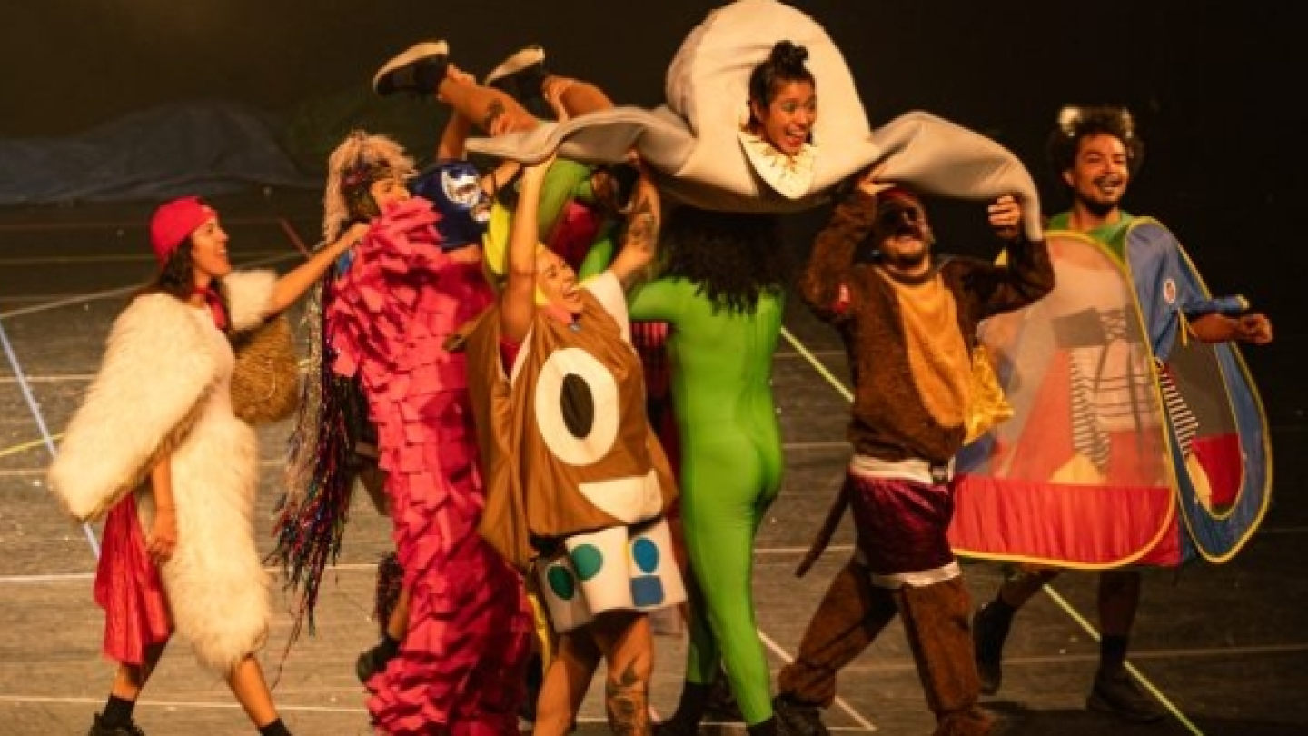 Fotografia de uma apresentação teatral com forte caráter lúdico e performático. No palco, vários artistas vestem figurinos coloridos e fantasiosos, representando personagens e criaturas estilizadas. Um dos intérpretes é erguido acima do grupo, enquanto os demais interagem em uma cena marcada por movimento, alegria e expressividade corporal.