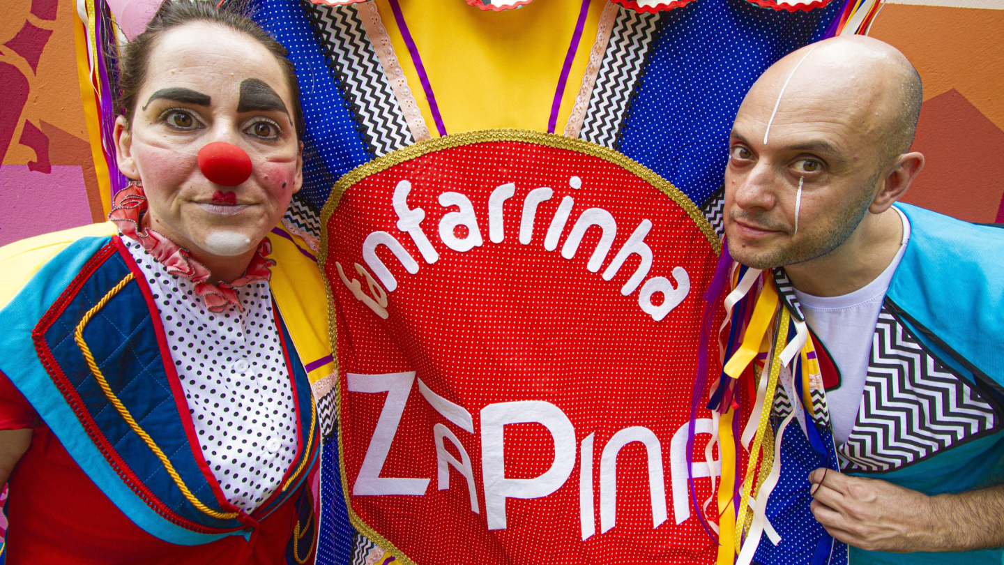 Dois artistas fantasiados e maquiados posicionados nas laterais de um estandarte colorido onde se lê "Fanfarrinha Za Pina"