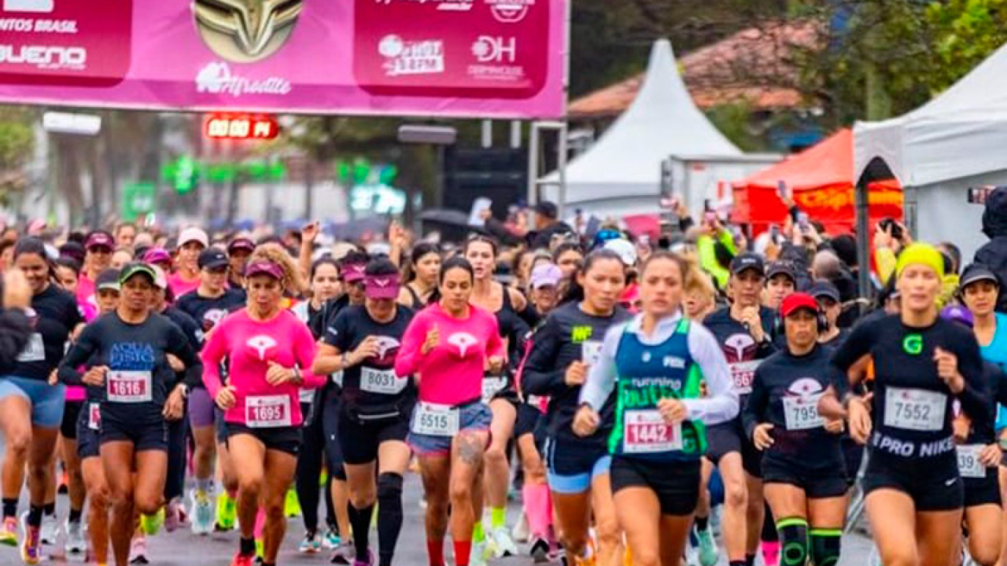Grande grupo de mulheres participa de uma corrida de rua organizada, passando sob um pórtico inflável rosa de largada ou chegada
