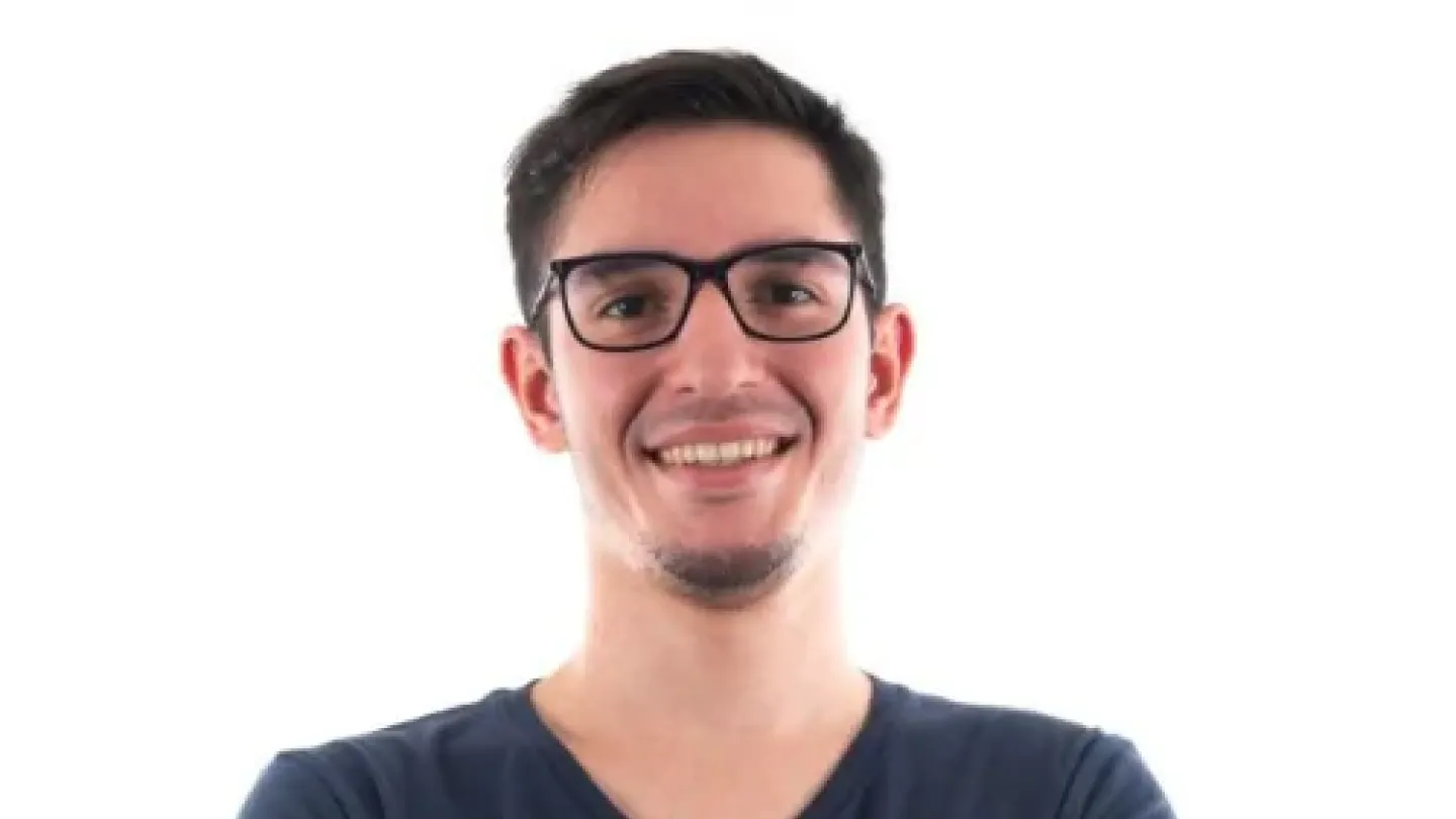 Fotografia do humorista Deco Machado. Ele aparece sorrindo, usando óculos de armação preta e camiseta azul escura, com os braços cruzados diante de um fundo claro. A imagem tem estilo de retrato profissional e destaca sua expressão amigável e descontraída.