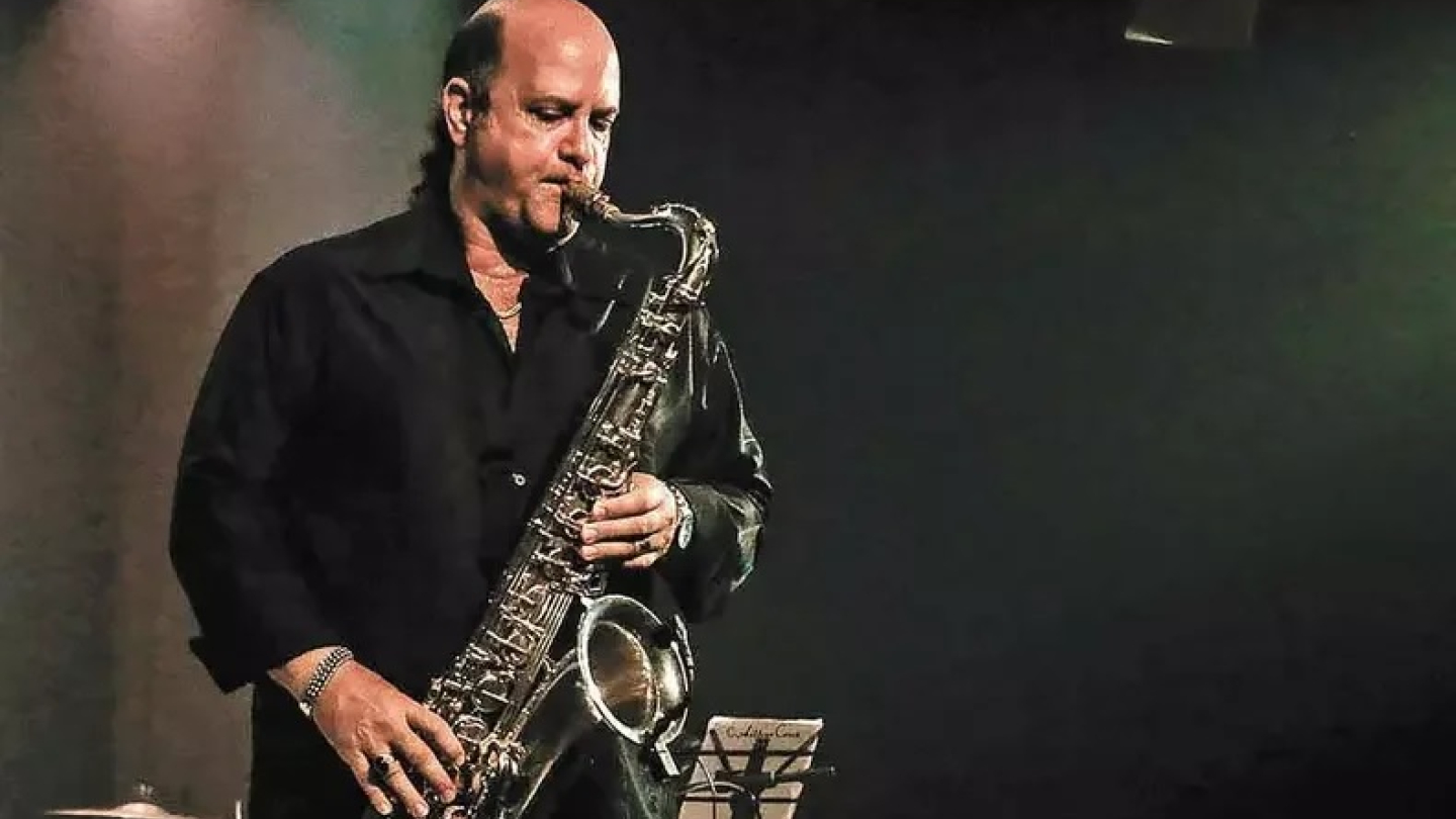 Um músico calvo vestido com uma camisa preta se apresenta concentrado no palco enquanto toca um saxofone