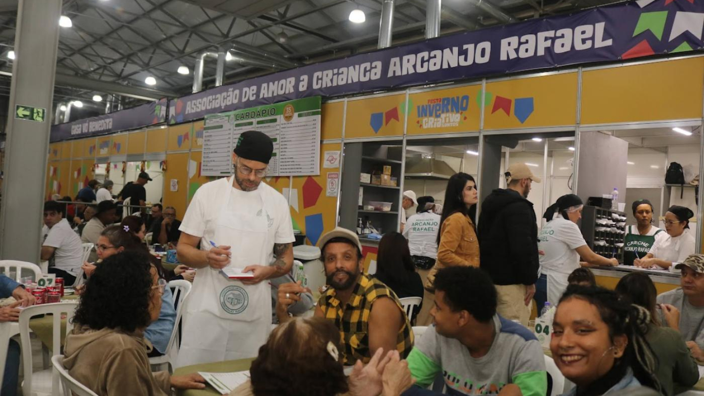 Garçom anotando o pedido de pessoas sentadas ao redor de uma mesa em um evento movimentado, com a barraca da "Associação de Amor à Criança Arcanjo Rafael" ao fundo
