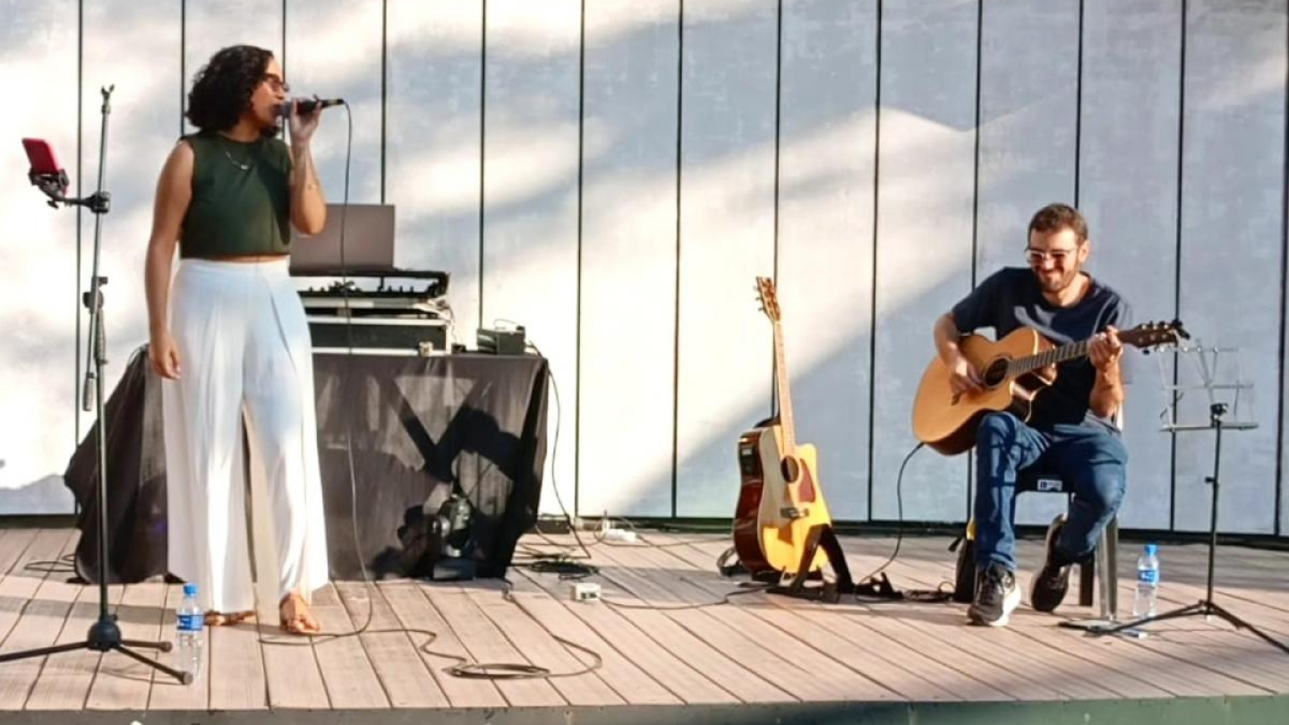 Uma cantora se apresenta em um palco iluminado ao lado de um músico sentado que a acompanha tocando violão
