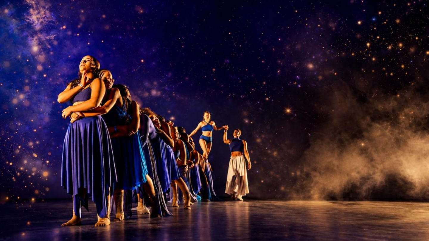 Grupo de bailarinos se apresentando no palco com figurinos em tons de azul e branco, alinhados sob um impressionante fundo cenográfico que simula um céu estrelado e uma galáxia iluminada