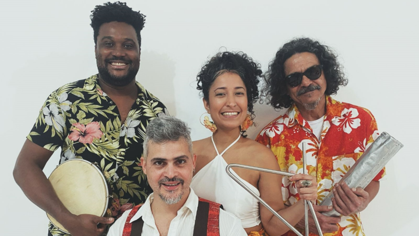 Fotografia promocional do grupo Adriano Salhab & Forró Xique-Xique. Quatro músicos posam diante de um fundo branco, usando roupas coloridas inspiradas na cultura popular nordestina. Os integrantes seguram instrumentos típicos como pandeiro, triângulo e ganzá, enquanto sorriem para a câmera. A imagem transmite um clima festivo e remete às tradições do forró e dos festejos populares brasileiros.