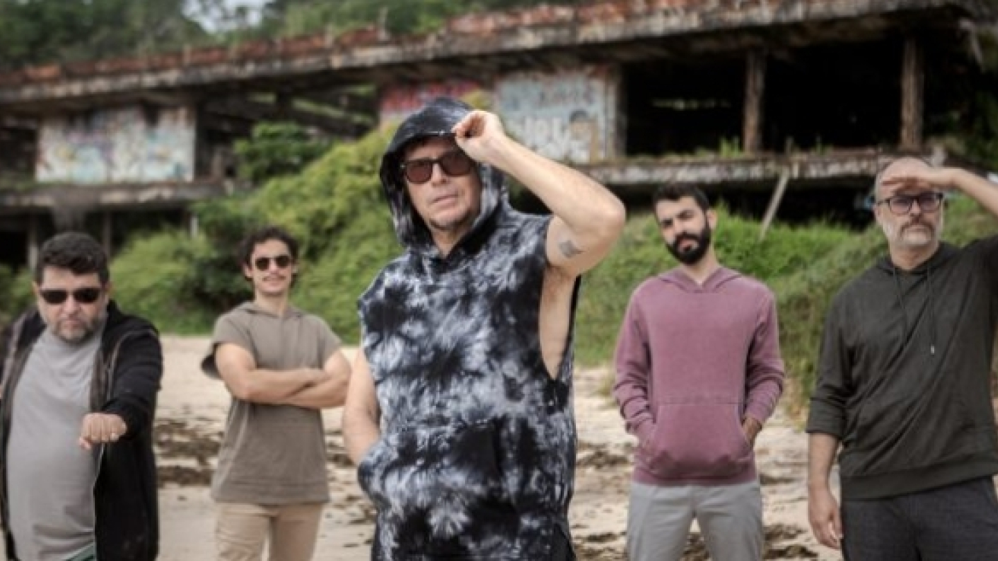 Imagem de divulgação da banda Mundo Livre S/A. Os cinco integrantes aparecem espalhados pela faixa de areia, tendo ao fundo uma estrutura antiga em ruínas parcialmente coberta pela vegetação. O músico central ocupa posição de destaque, usando óculos escuros e uma peça com capuz em tons de preto e cinza, enquanto os demais integrantes vestem roupas casuais em cores neutras. A combinação entre o cenário litorâneo, a arquitetura decadente e a postura descontraída dos músicos transmite uma estética urbana e alt