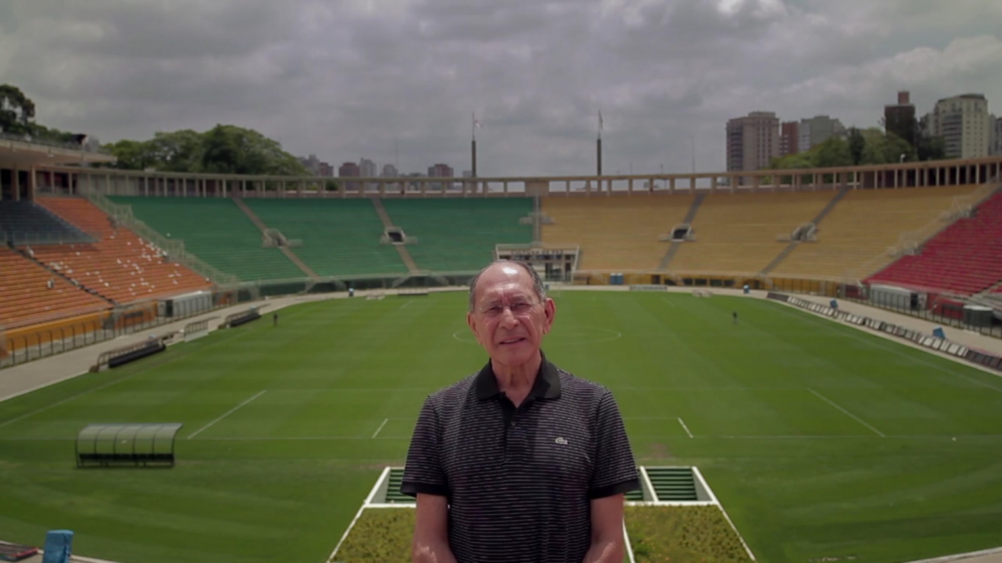 Cena do documentário "O Pai do Gol" mostrando José Silvério em primeiro plano, usando camisa polo escura, posicionado à beira do campo de um grande estádio de futebol vazio. Ao fundo aparecem o gramado, arquibancadas nas cores verde, amarelo, vermelho e laranja, além de prédios da cidade sob um céu parcialmente nublado.