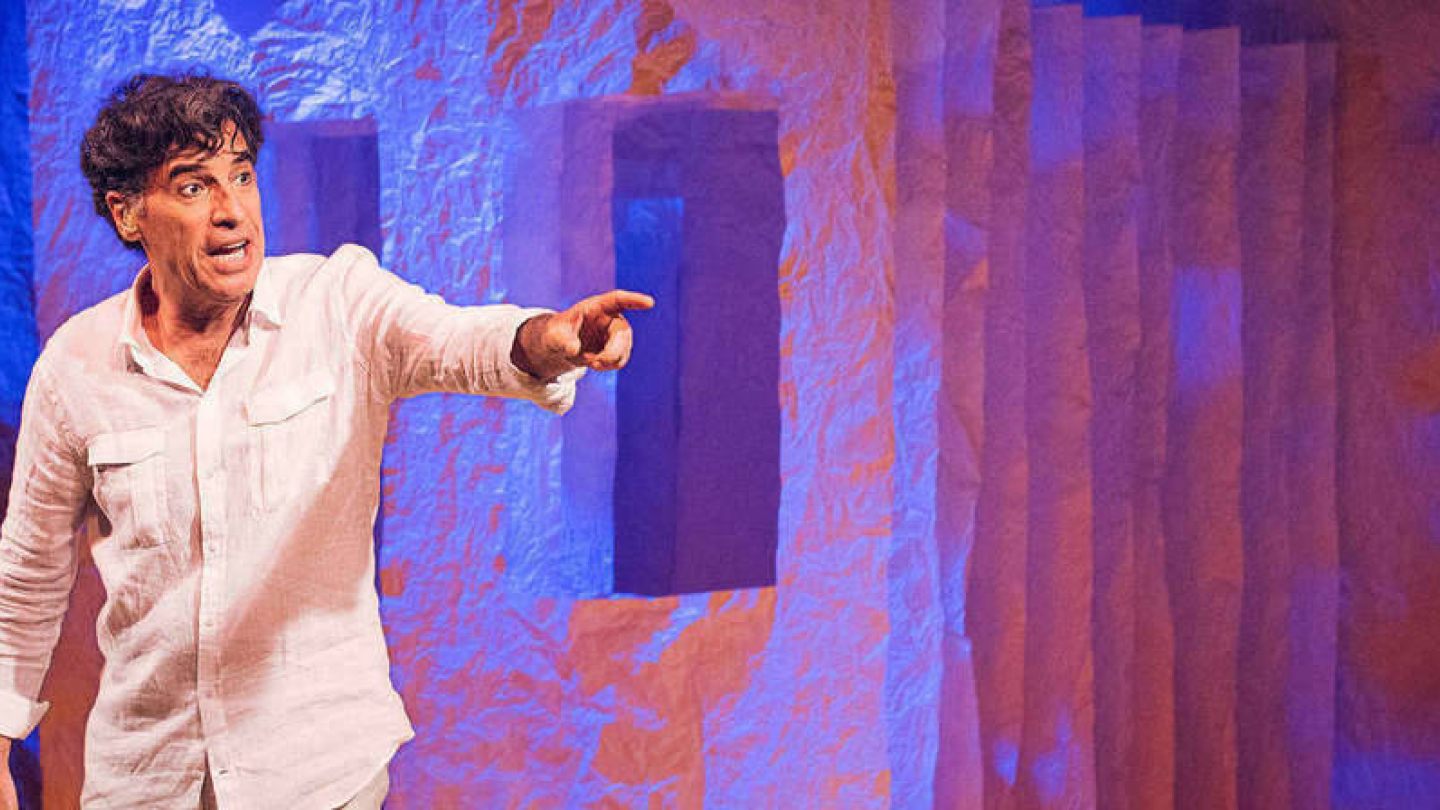 Ator Paulo Betti em um palco de teatro, vestindo uma camisa branca e apontando para frente com uma expressão expressiva, diante de um cenário com texturas iluminadas em tons de azul e laranja