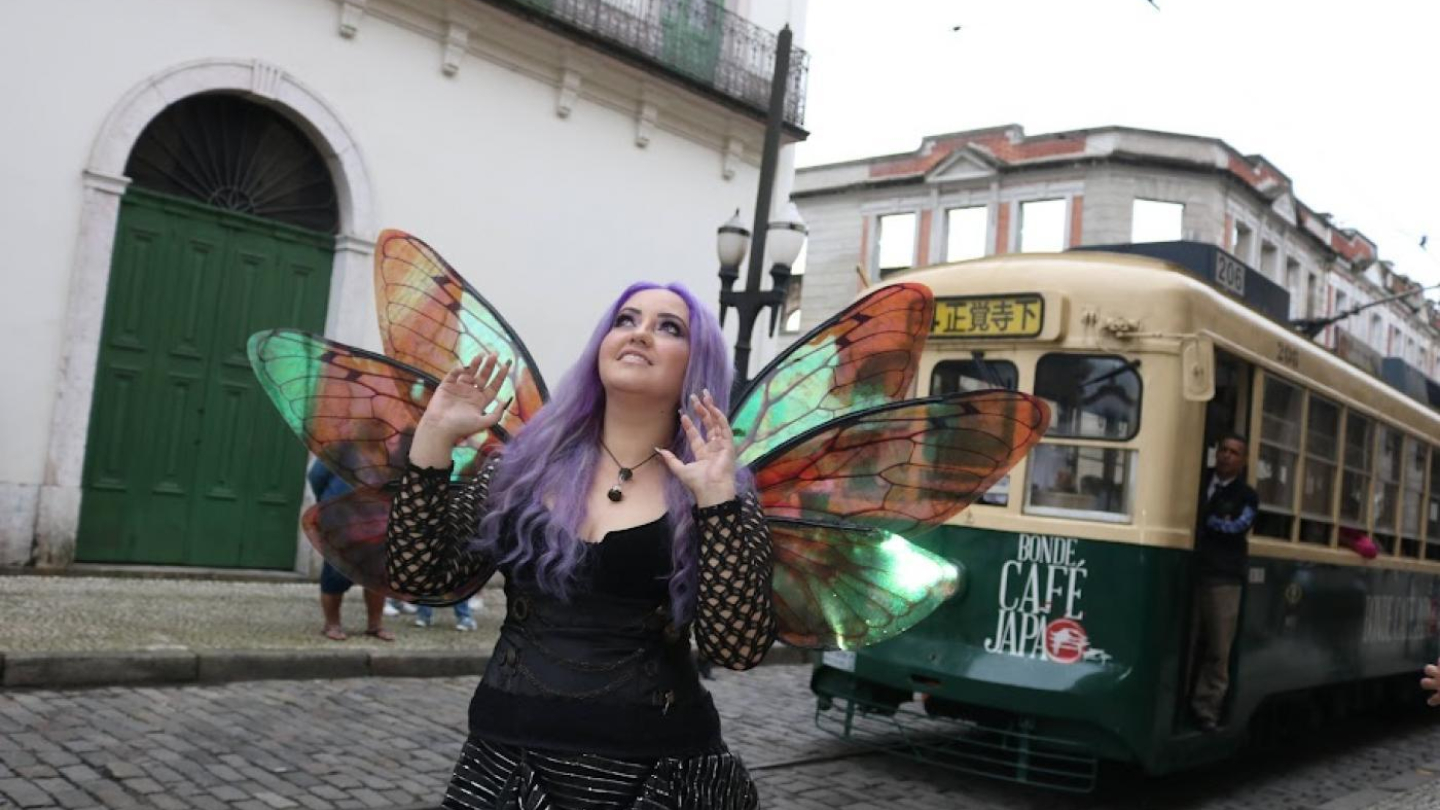 Mulher de cabelos roxos e asas de fada iridescentes posando em uma rua de paralelepípedos, com um bonde histórico ao fundo