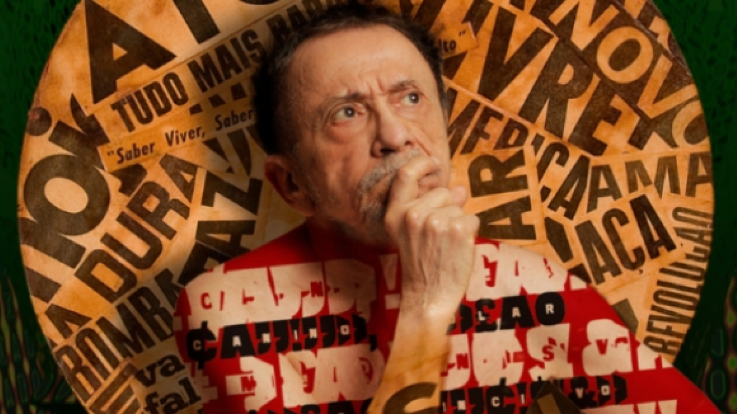 Fotografia promocional do cantor e compositor Tom Zé. O artista aparece em primeiro plano com expressão contemplativa, apoiando a mão no rosto como se estivesse refletindo ou criando uma nova ideia. Atrás dele, um grande painel circular reúne palavras e fragmentos de texto sobrepostos, criando uma colagem visual característica da estética experimental associada à sua obra. A combinação de tons quentes, o fundo verde e a tipografia espalhada pela imagem reforçam a sensação de criatividade, inquietação intele