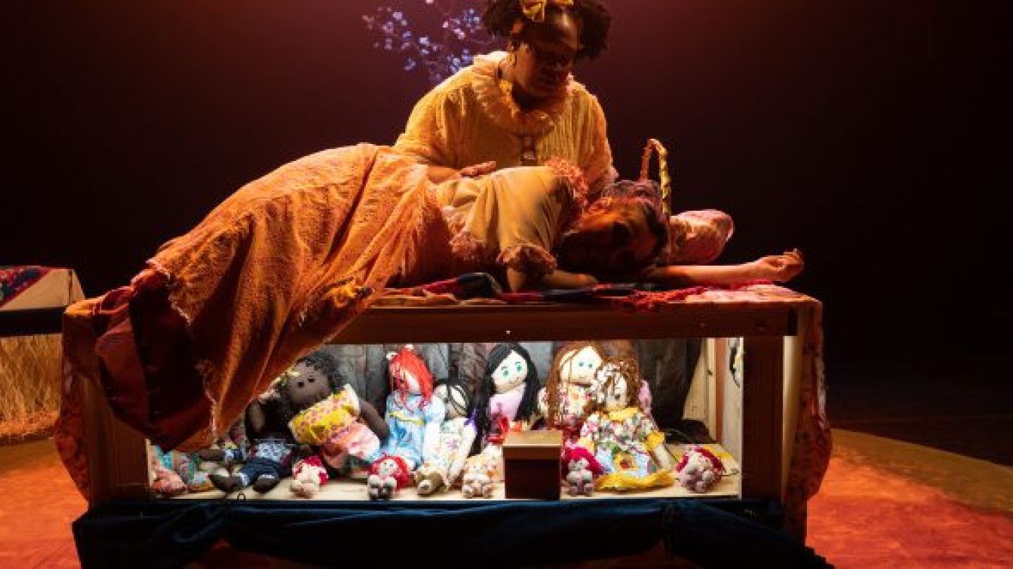 Cena teatral iluminada por luzes quentes, mostrando uma mulher sentada ao lado de uma criança deitada sobre uma cama de madeira, em um momento de acolhimento e carinho. Sob a cama, diversas bonecas de pano coloridas ficam expostas, compondo o cenário. Ao fundo, o palco escuro destaca os personagens e reforça a atmosfera sensível e poética da apresentação.