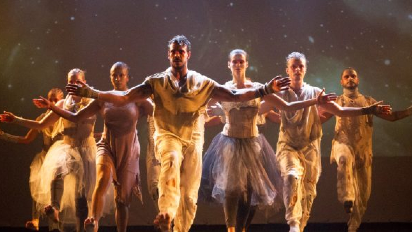 Sete bailarinos ocupam o palco em formação, com os braços abertos e um deles à frente do grupo. Vestidos com figurinos em tons claros, eles são iluminados por luz quente diante de um fundo escuro, transmitindo força, unidade e expressividade corporal.
