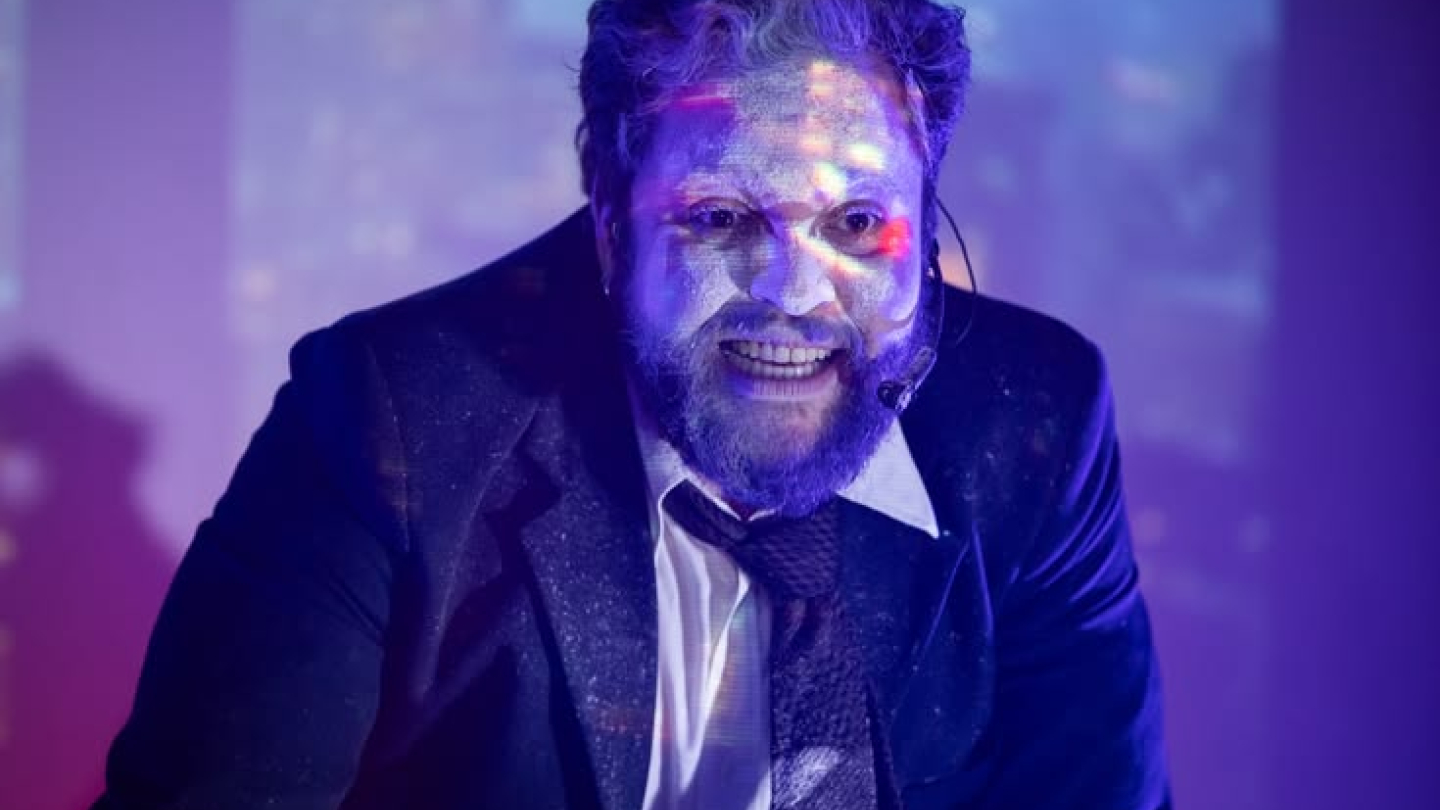 Um ator aparece em primeiro plano usando terno escuro, camisa branca e gravata, com o rosto coberto por tinta branca e iluminado por luzes em tons de azul e roxo. Sua expressão intensa e o uso de microfone de cabeça reforçam o caráter performático da cena.