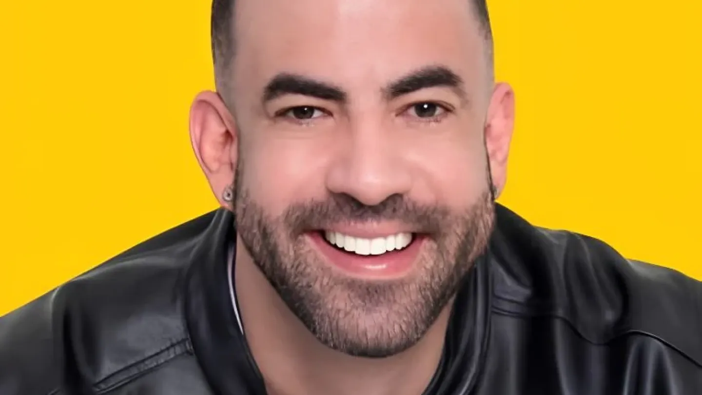 Foto de divulgação de um homem sorridente, com barba aparada e cabelo curto escuro, usando uma jaqueta preta sobre camiseta branca. O fundo é amarelo sólido, destacando seu rosto e expressão simpática.