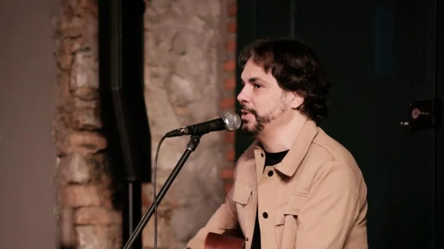 Um cantor se apresenta sentado, tocando violão e cantando ao microfone. Ele veste jaqueta bege sobre camiseta escura, e o palco tem ao fundo uma parede de tijolos aparentes, criando um ambiente intimista.