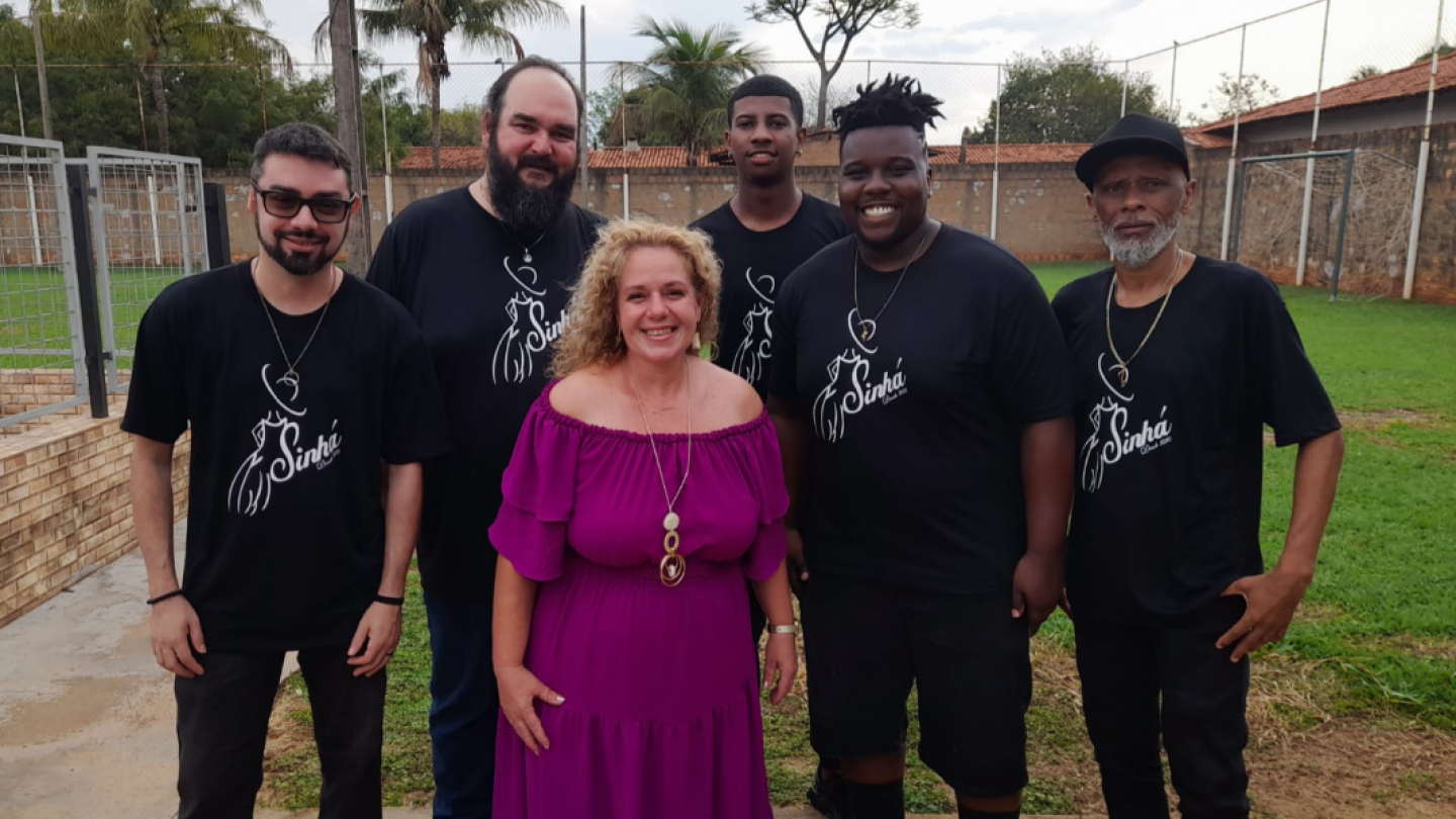 oto promocional de um grupo formado por seis homens e uma mulher, reunidos ao ar livre e sorrindo para a câmera. A mulher veste um vestido rosa, enquanto os demais usam camisetas pretas com o logotipo do grupo, em um ambiente com gramado e quadra esportiva ao fundo.