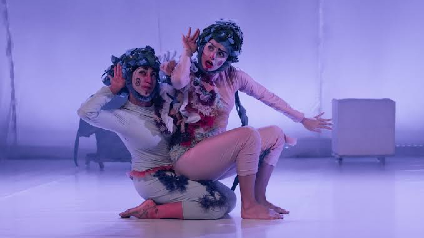Dois performers estão no palco em uma pose expressiva, usando figurinos em tons claros com aplicações de tecido e maquiagem cênica. Sob iluminação em tons de lilás, ambos gesticulam de forma teatral, criando uma cena que remete a personagens fantásticos ou surreais.