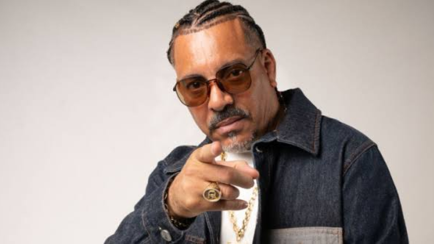 Foto promocional do rapper Dexter em primeiro plano, usando óculos escuros, correntes e jaqueta jeans. Com expressão séria, ele aponta para a câmera, reforçando a presença marcante e a postura firme que caracterizam sua trajetória no rap nacional.