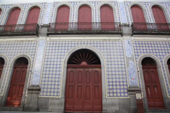 A fachada de um prédio histórico de dois andares, com a parte de baixo coberta por azulejos brancos e azuis. No andar superior, uma sacada com corrimão de ferro forjado e janelas de madeira avermelhada. No andar de baixo, portas de madeira vermelha com formato de arcos