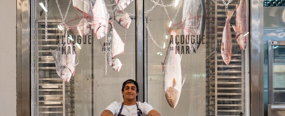 Homem com avental e boné sorri sentado em frente a uma vitrine refrigerada com grandes peixes pendurados, no box “Açougue do Mar”