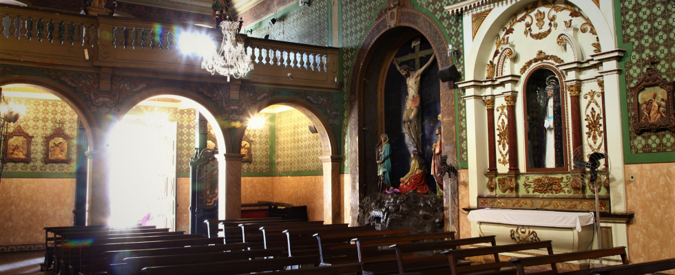 Interior de uma igreja antiga com bancos de madeira, arcos e paredes ricamente decoradas. Há um crucifixo e uma estátua de santa em nichos, e a luz do sol entra forte por uma porta aberta à esquerda