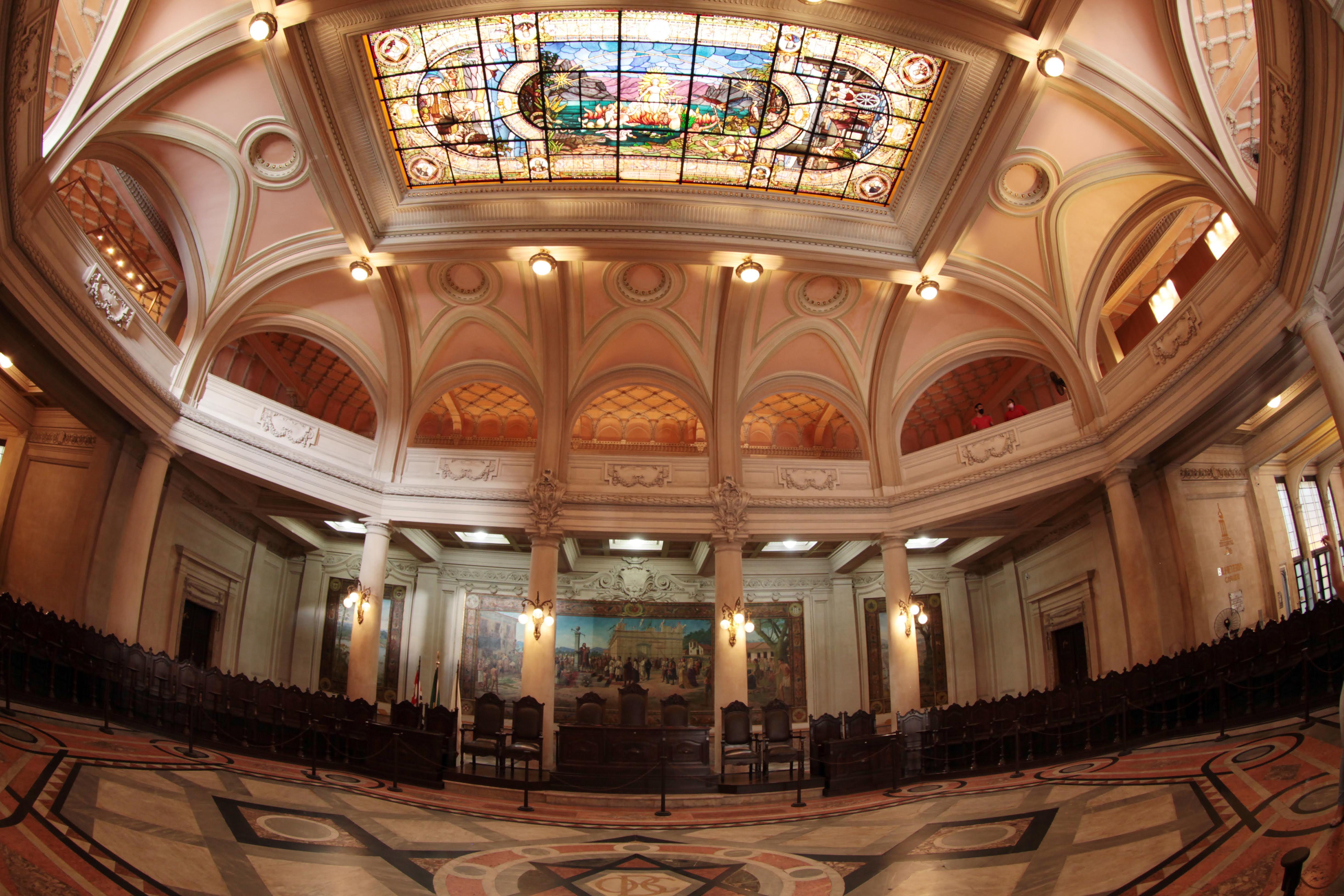 Salão nobre histórico com vitral colorido no teto, arcos ornamentados e cadeiras de madeira dispostas ao redor