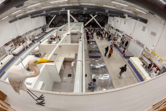 Vista ampla do interior do Mercado de Peixes de Santos, com corredores de boxes, clientes circulando e, em primeiro plano, uma escultura de garça branca com bico amarelo.