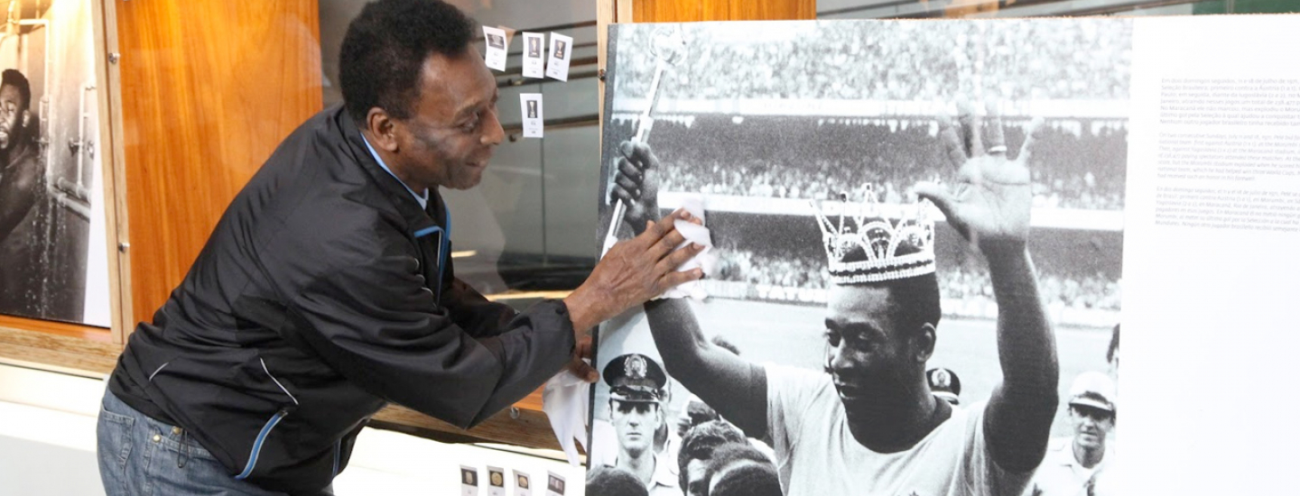 Pelé sorri enquanto toca uma foto em preto e branco de si mesmo, coroado em campo diante da torcida