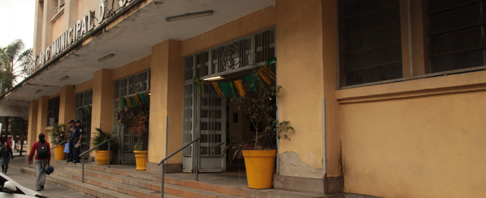 Fachada do Mercado Municipal de Santos, em prédio amarelo com escadaria de acesso, vasos grandes com plantas e pessoas entrando e saindo