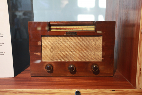 Rádio antigo de madeira, com três botões frontais e tela de tecido bege, exposto em vitrine de museu.