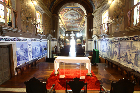 Interior de uma igreja ou capela com bancos de madeira alinhados, paredes adornadas com azulejos azuis e brancos, e pinturas no alto. Há um altar coberto de branco no centro, com tapete vermelho e duas cadeiras escuras na frente, e luz forte vindo do fundo