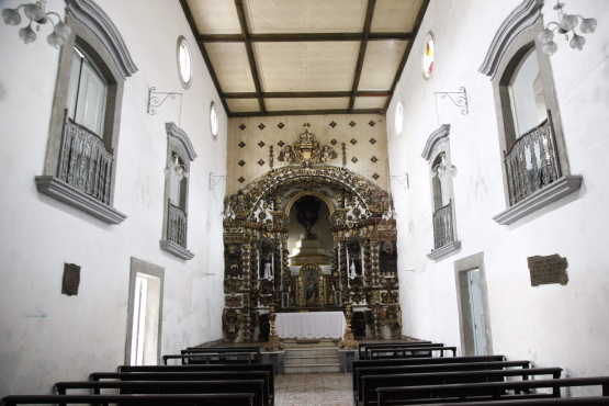 Interior de uma capela rústica, com paredes brancas lisas e um teto de madeira com vigas. Ao fundo, um altar barroco escuro e ornamentado domina a cena. Bancos de madeira simples ocupam a nave, e há janelas com pequenas sacadas de ferro forjado e algumas coloridas nas laterais