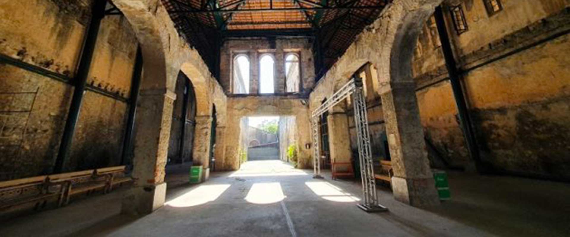 O interior de um prédio em ruínas, com paredes de pedra e cimento. Há arcos e colunas que sustentam o telhado, e um corredor longo leva à luz que entra pela saída. O sol cria sombras no chão