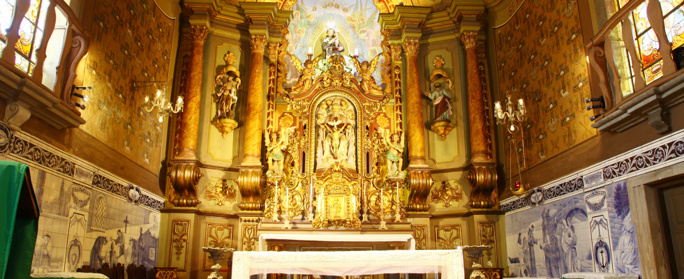 Interior de uma capela ricamente decorada, com um altar-mor dourado e ornamentado no centro, ladeado por colunas e estátuas. As paredes laterais possuem azulejos azuis e brancos na parte inferior e pinturas coloridas acima, com janelas de vitral visíveis