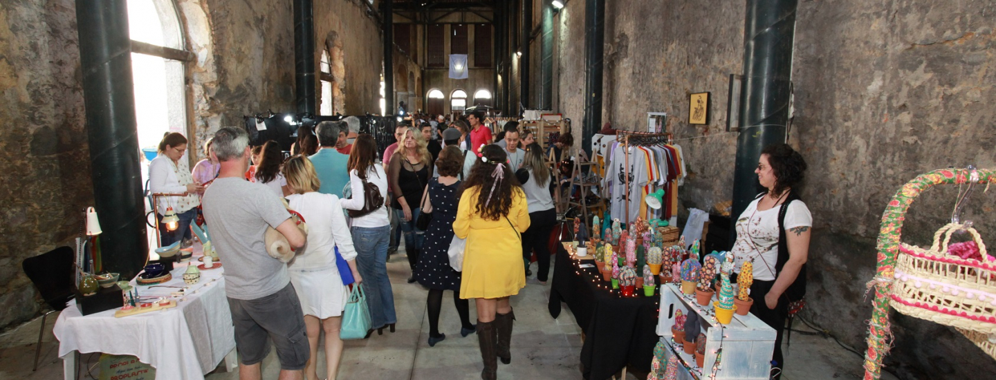 Um salão grande, com paredes e colunas de pedra desgastada, onde acontece uma feira de artesanato. Muitas pessoas circulam entre as barracas com produtos variados, como cerâmica e bonecos