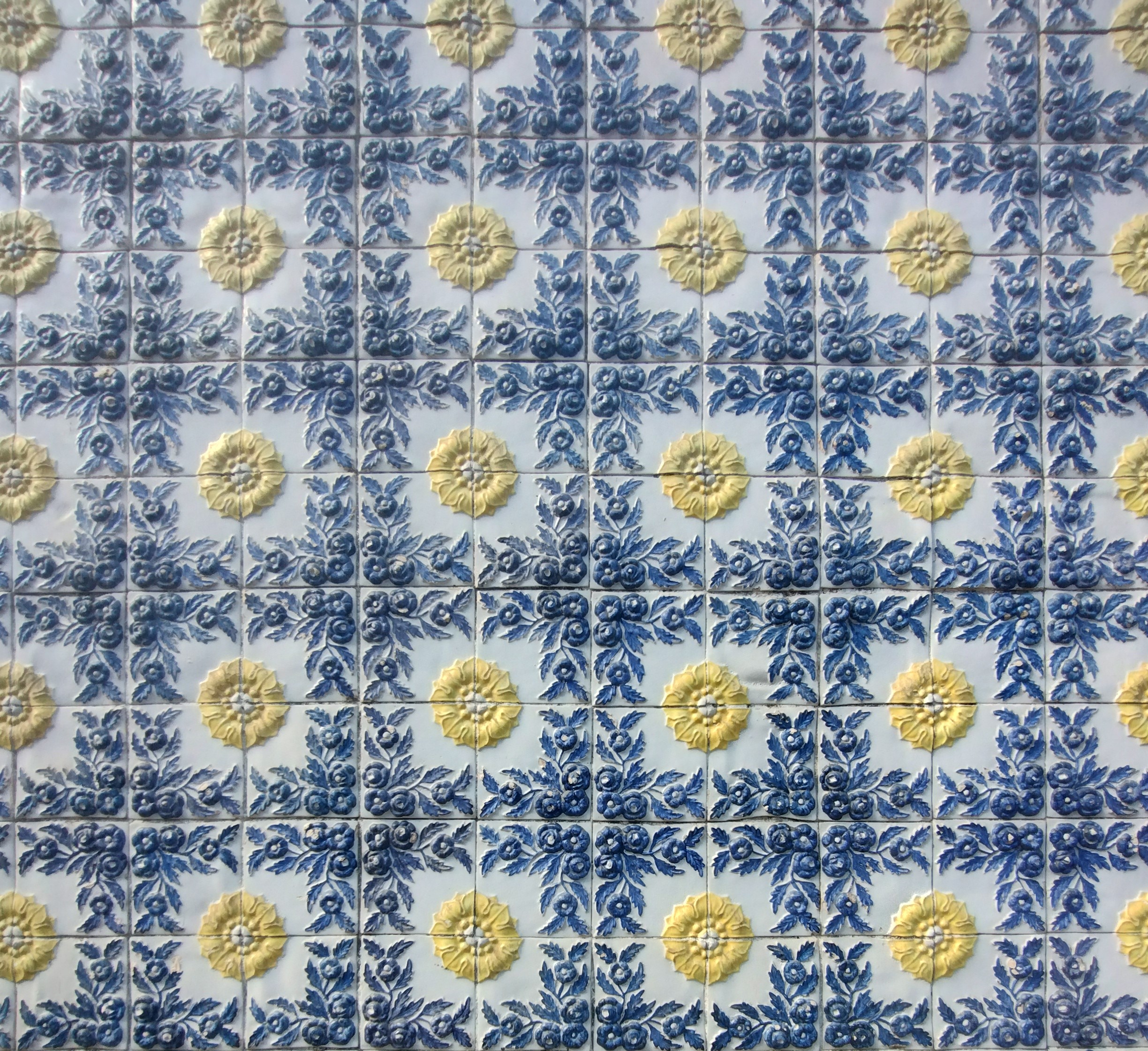 Uma parede de azulejos antigos com padrões florais. Em fundo branco, os azulejos alternam entre fileiras de desenhos de flores azuis e folhagens, e fileiras de grandes flores amarelas