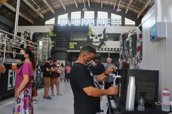 Pessoas circulam e bebem chope em um galpão da cervejaria Everbrew, decorado com murais em preto e branco e grandes tanques de produção ao fundo