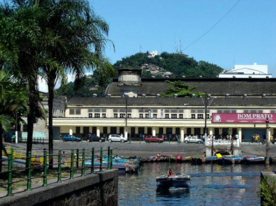 Vista externa do Mercado Municipal de Santos, em frente ao canal com barcos atracados. Ao fundo, o Monte Serrat com o santuário no topo