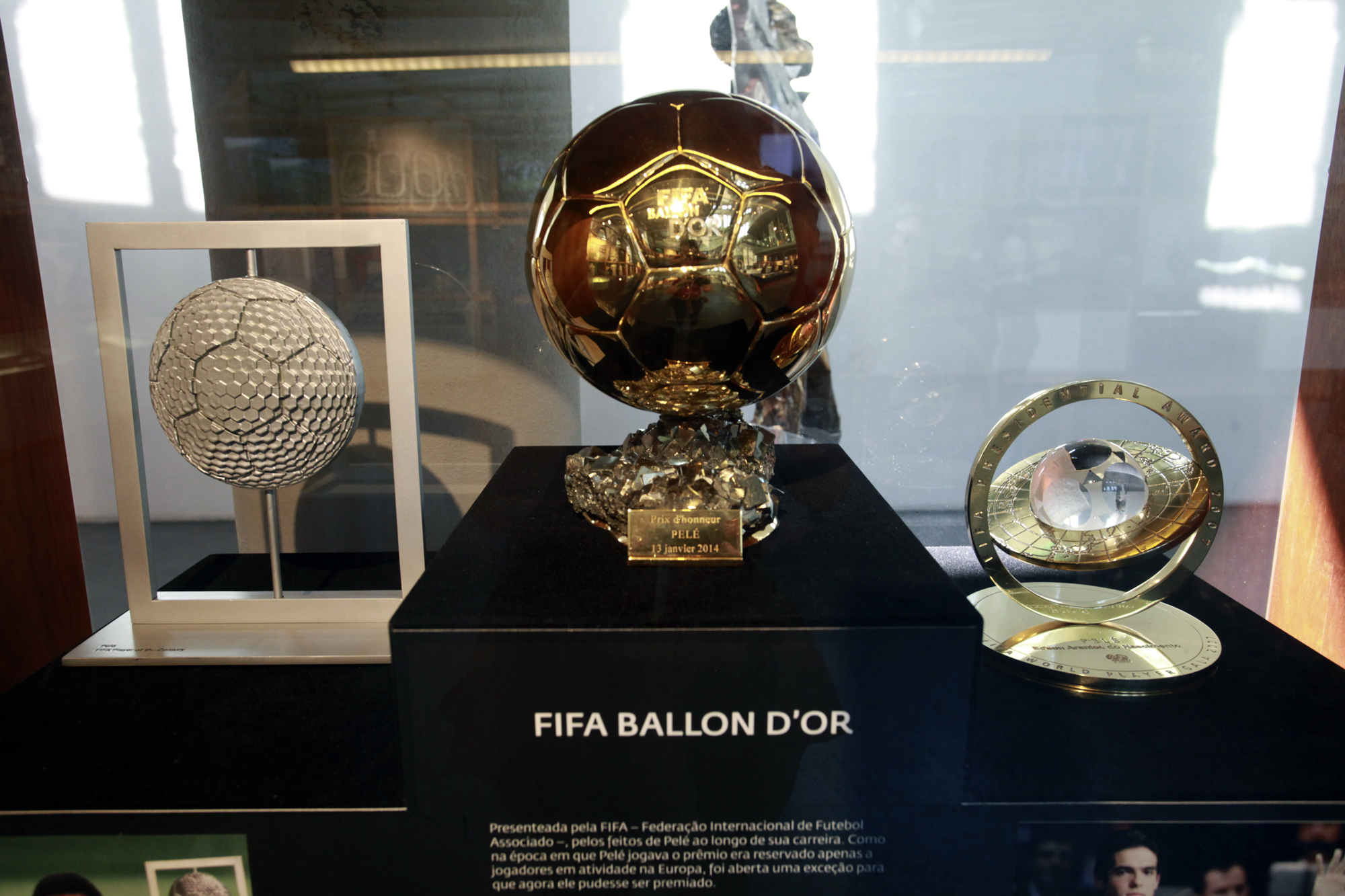 Três troféus em exposição: ao centro, a Bola de Ouro da FIFA em dourado; à esquerda, escultura prateada em formato de bola; e à direita, prêmio circular dourado com esfera de cristal no centro.