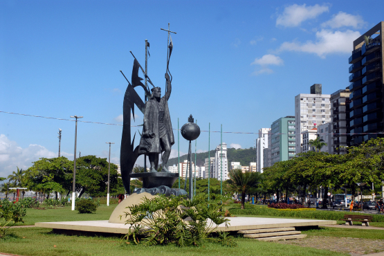 A imagem mostra o monumento a Brás Cubas, fundador da cidade de Santos, localizado na orla da praia. A escultura em bronze retrata Brás Cubas erguendo uma cruz em uma das mãos e um estandarte na outra, simbolizando a fé e a fundação da cidade. Ao redor, há um amplo gramado com jardins bem cuidados e, ao fundo, edifícios residenciais e comerciais que compõem a paisagem urbana santista. O céu azul e o clima ensolarado reforçam a atmosfera praiana e vibrante do cenário, que une história, arte e modernidade.
