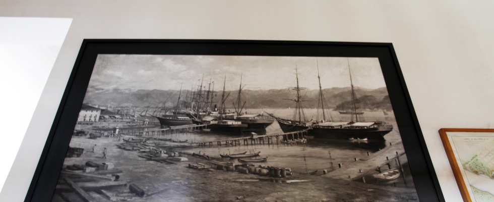 Quadro em preto e branco retratando um antigo porto com vários navios atracados, pequenas embarcações e barris espalhados pelo cais. Ao fundo, montanhas e céu nublado.