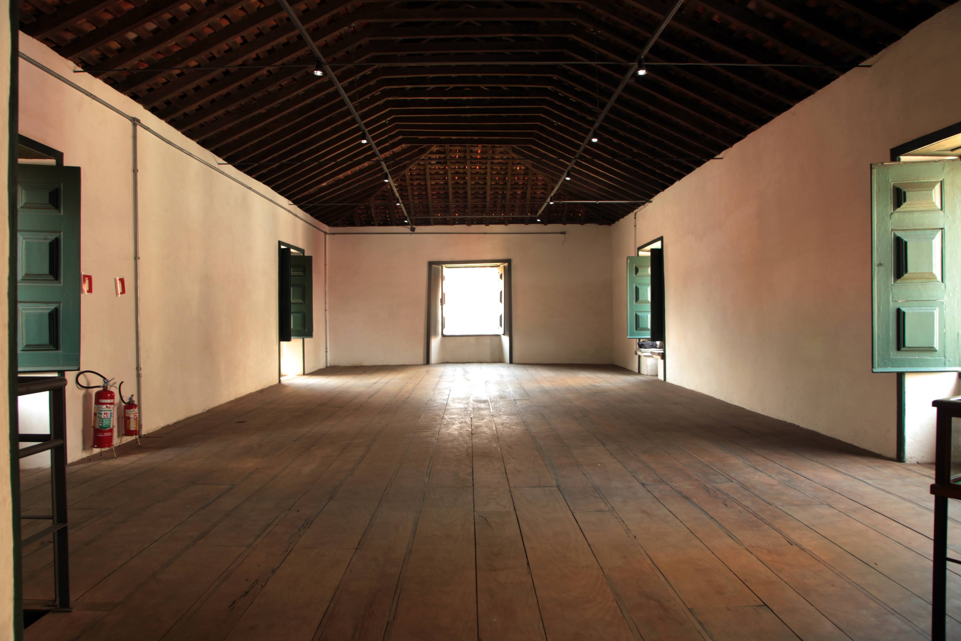 Sala ampla e vazia com piso e teto de madeira, janelas com venezianas verdes e iluminação natural, em ambiente de estilo tradicional
