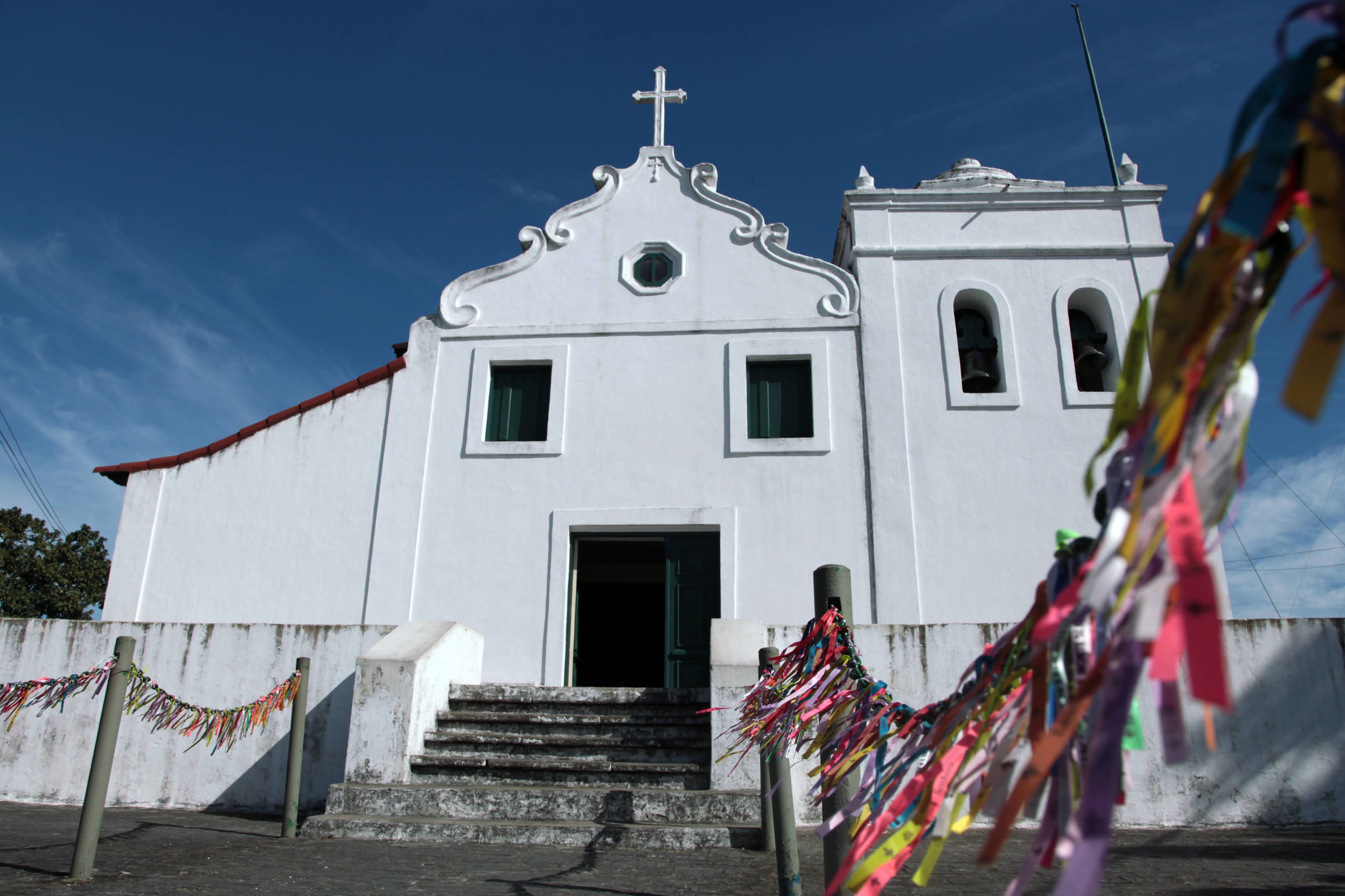 Igreja colonial branca no topo de escadaria, com torre de sinos e fitas coloridas decorando o caminho, sob céu azul claro