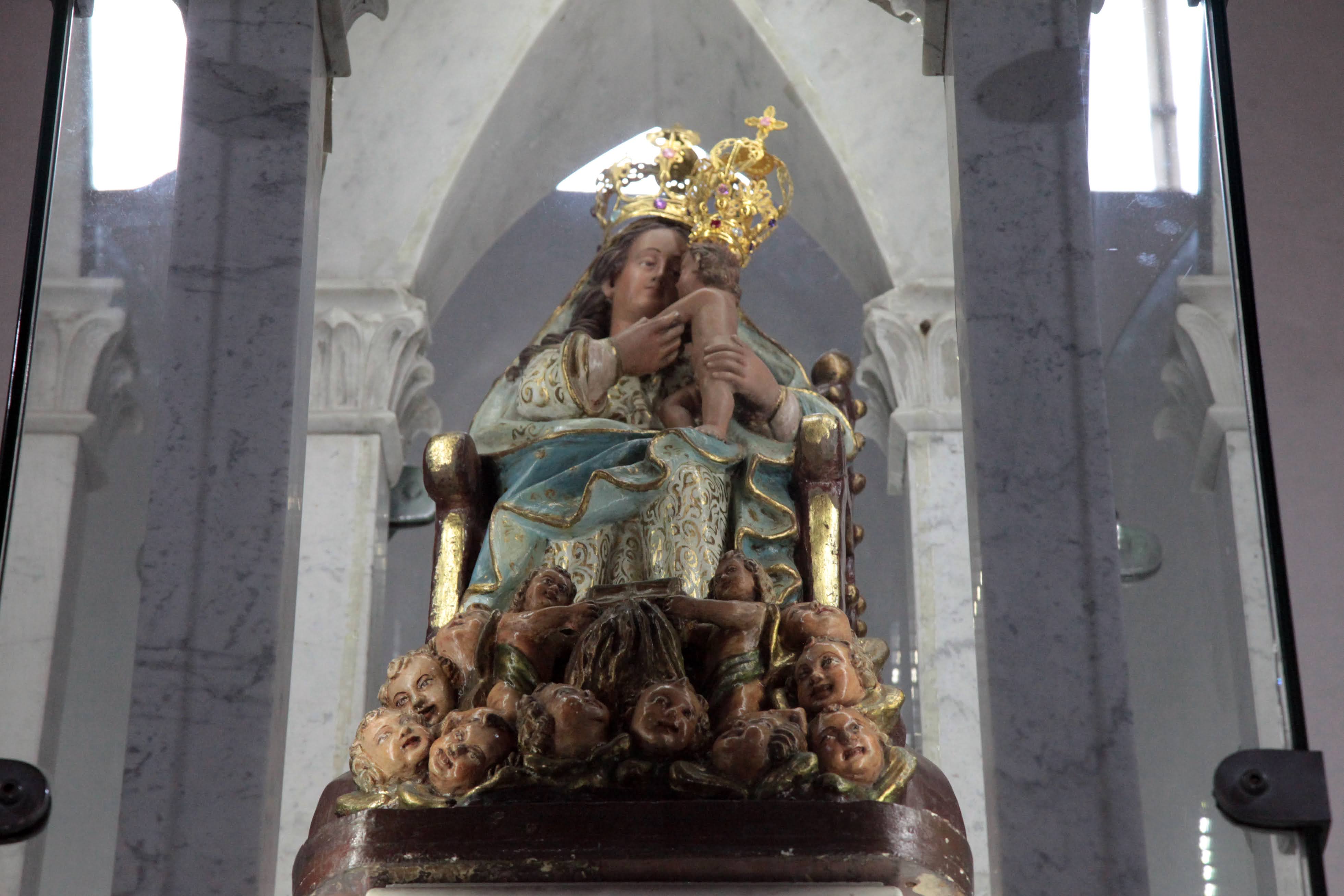 Imagem de Nossa Senhora entronizada com o Menino Jesus no colo, cercada por cabeças humanas esculpidas, em ambiente de igreja com arquitetura gótica
