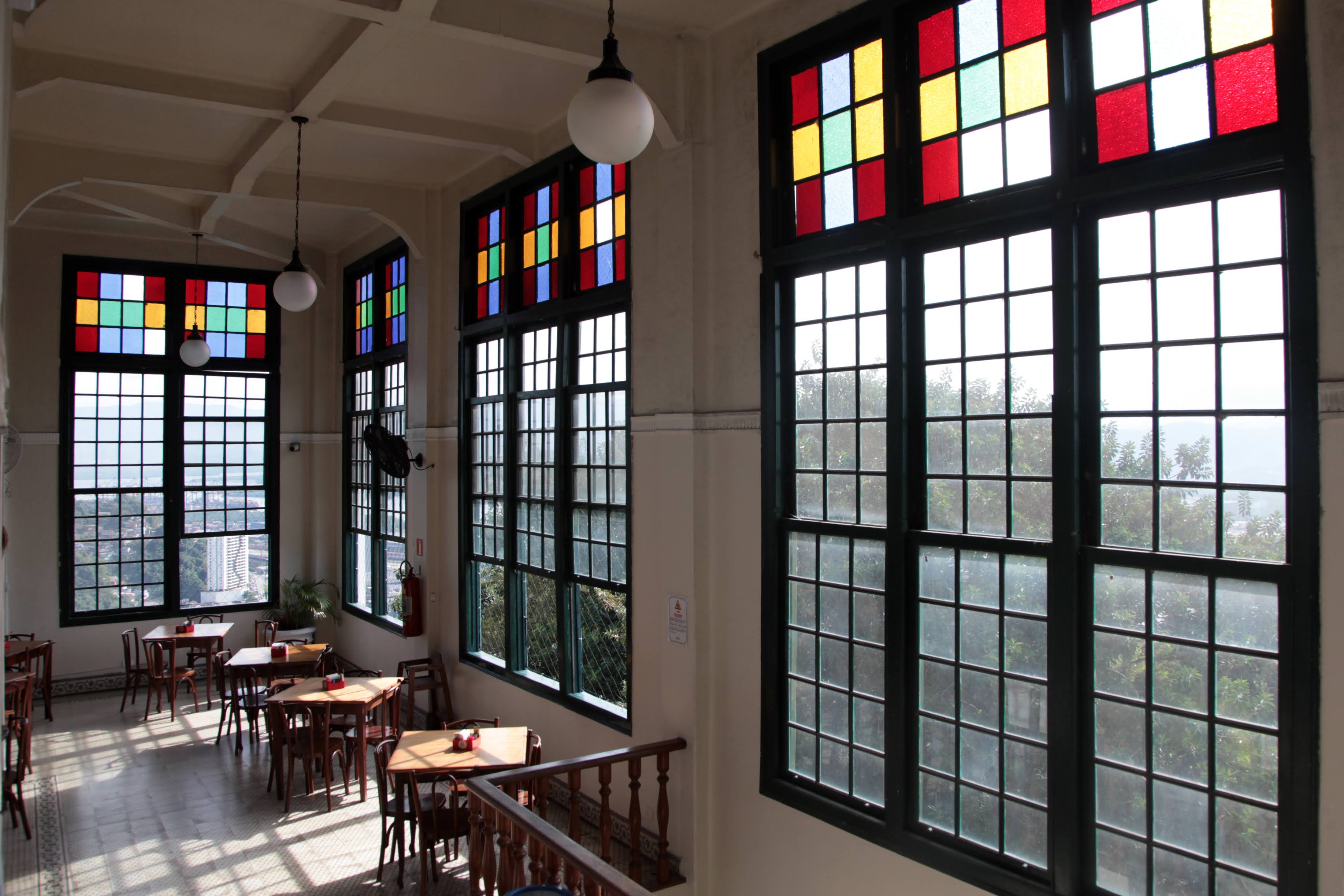 Sala iluminada com janelas grandes e vitrais coloridos, mobiliada com mesas e cadeiras de madeira, sugerindo um ambiente de café ou restaurante