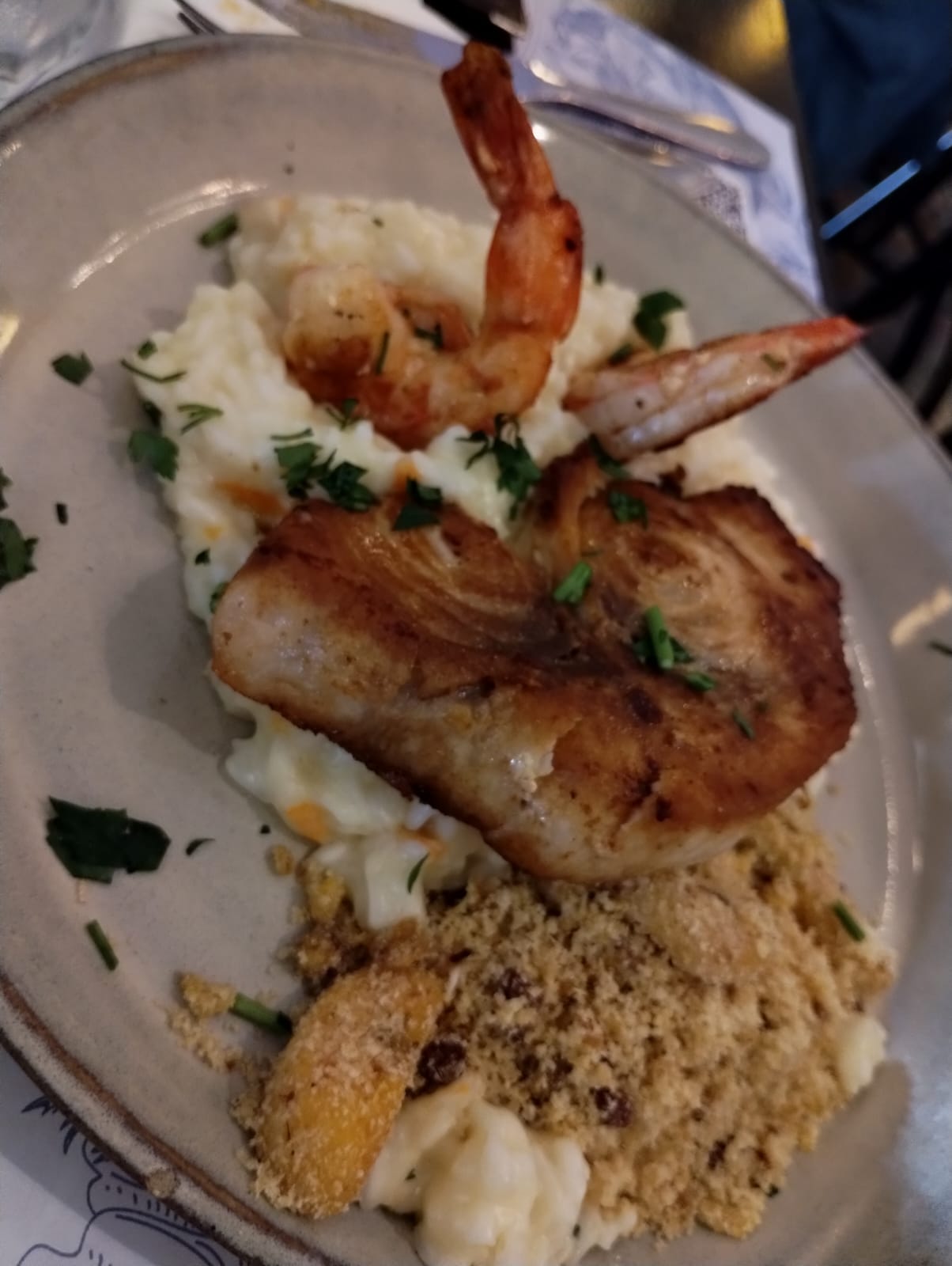 A imagem mostra um prato bem apresentado com peixe grelhado acompanhado de camarões. A proteína está disposta sobre uma porção cremosa de risoto. Ao lado, há uma porção de farofa dourada com pedaços de fruta, que parecem ser gomos de laranja, e possivelmente passas, acrescentando um toque agridoce ao prato.