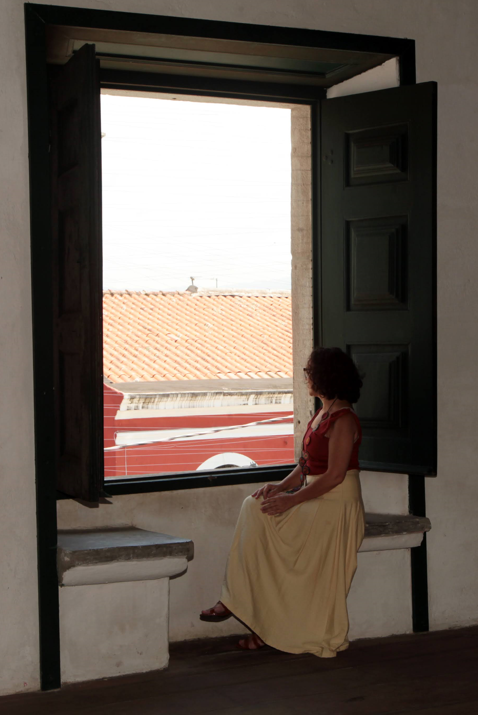 Mulher sentada em parapeito de janela, com roupa vermelha e saia bege, observa o exterior iluminado, criando uma cena contemplativa entre luz e sombra